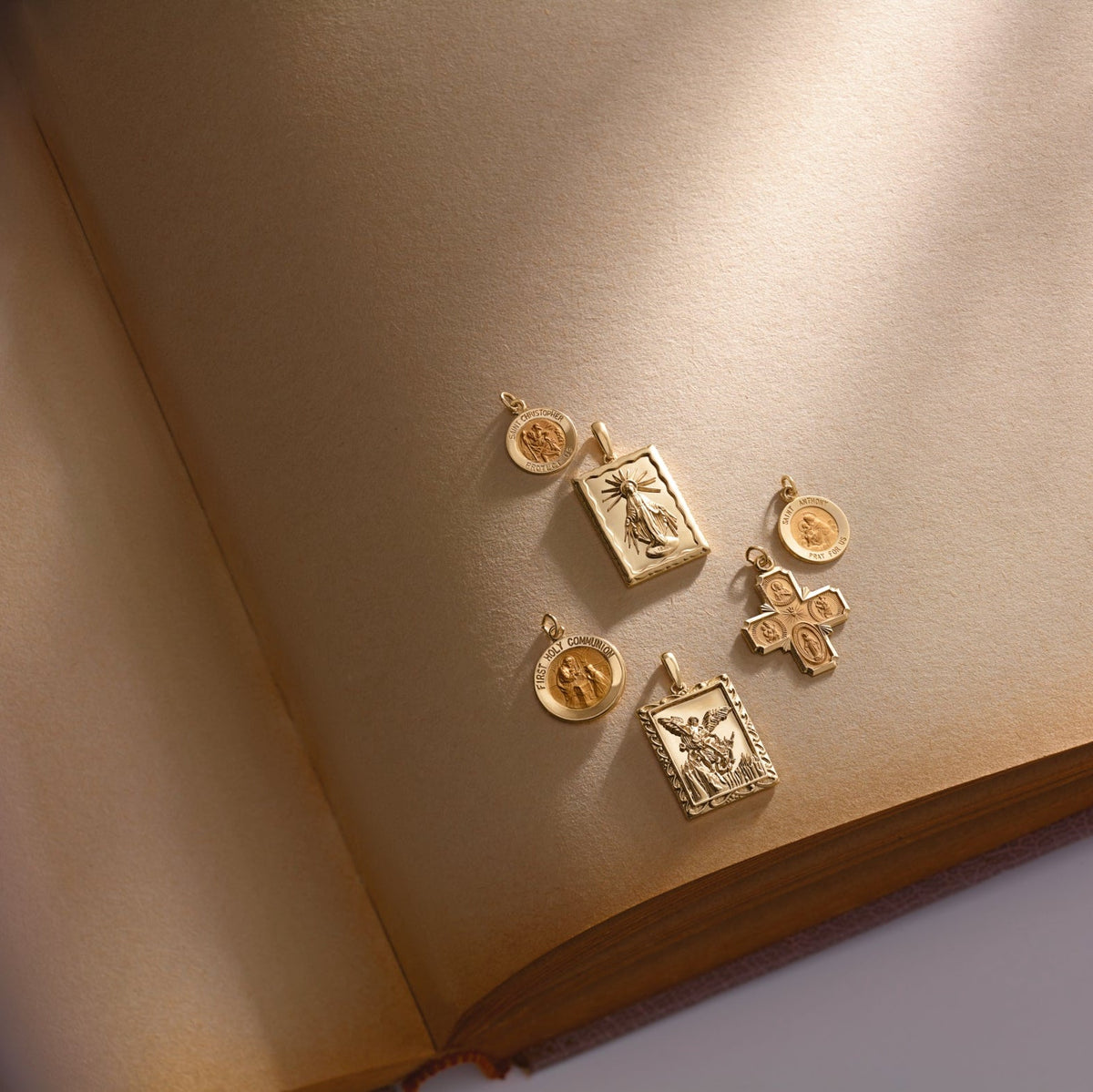 Assorted 14K gold religious charms resting on aged book pages, including saints, a cross, and First Holy Communion pendants.