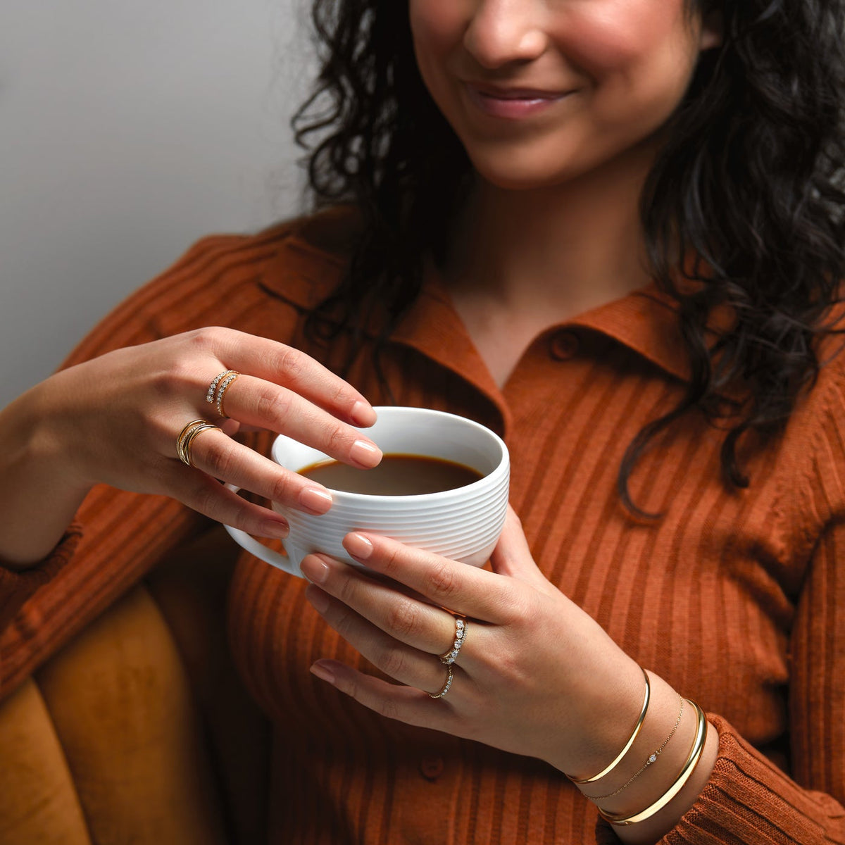 Woman holding coffee wearing elegant 14K gold rings with diamonds and matching gold bangles.